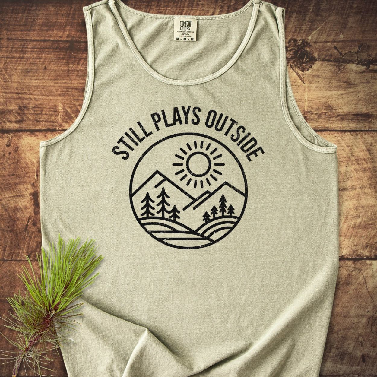 Still Plays Outside Tank Top, heather-gray with a black line-art graphic of a sun, mountains, and pine trees, laid flat on wood, styled with a pine sprig beside it.