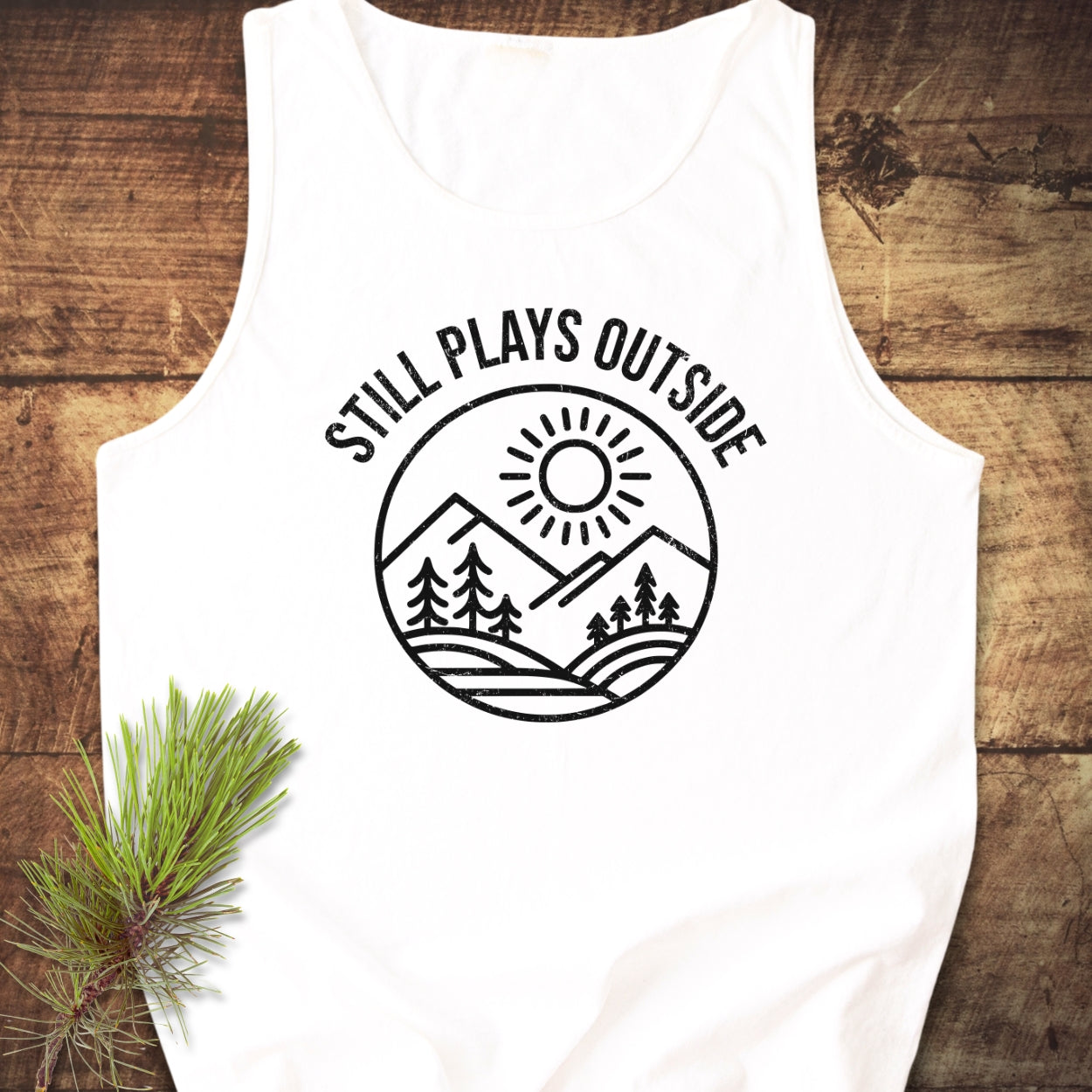 Still Plays Outside Tank Top features a round graphic of mountains, pine trees, and a sun with the slogan above, laid on wood, emphasizing a blend of comfort, style, and sustainability.