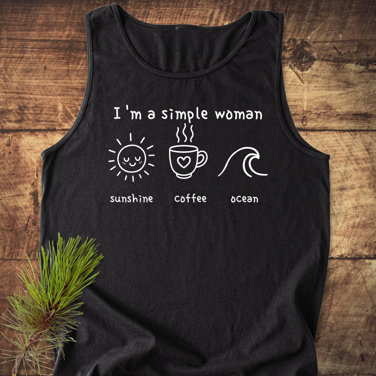 I'm A Simple Woman - Sunshine Coffee Ocean Tank Top featuring white print icons and text on a black shirt, laid on a wooden surface with a pine sprig, showcasing comfort and sustainability.