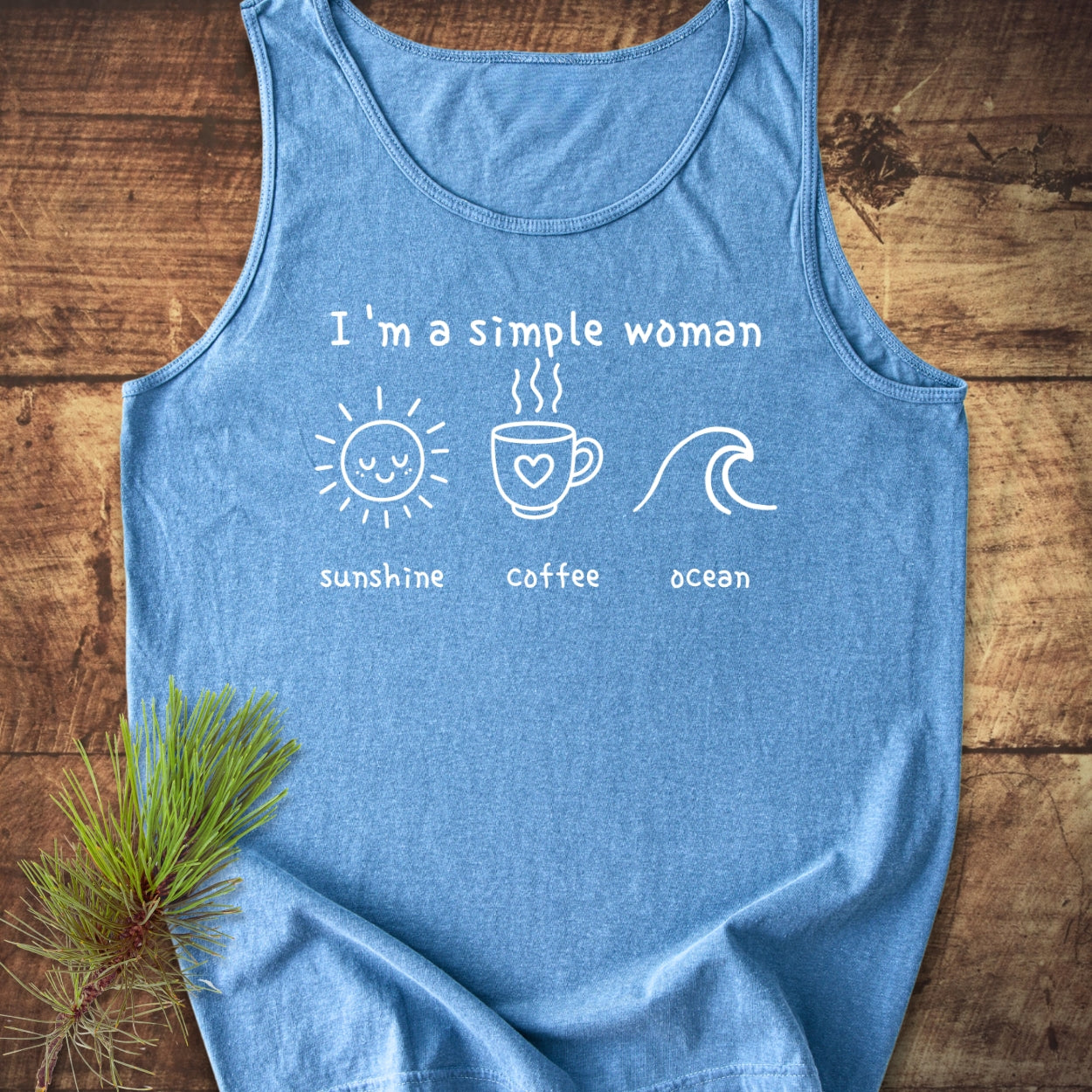 I'm A Simple Woman - Sunshine Coffee Ocean Tank Top features a heather-blue design with minimalist icons, showcasing love for sunshine, coffee, and ocean, laid on rustic wood with a pine branch accent.