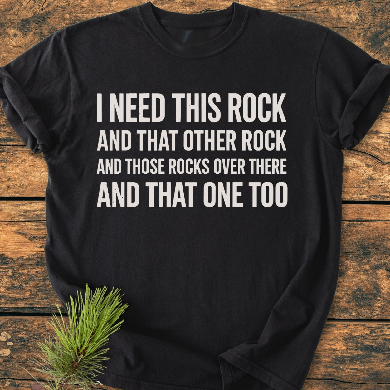 I Need This Rock - Vintage Wash Tee featuring bold white text on a black T-shirt, displayed on a wooden surface with a pine branch accenting the corner, exemplifying comfort and durability.