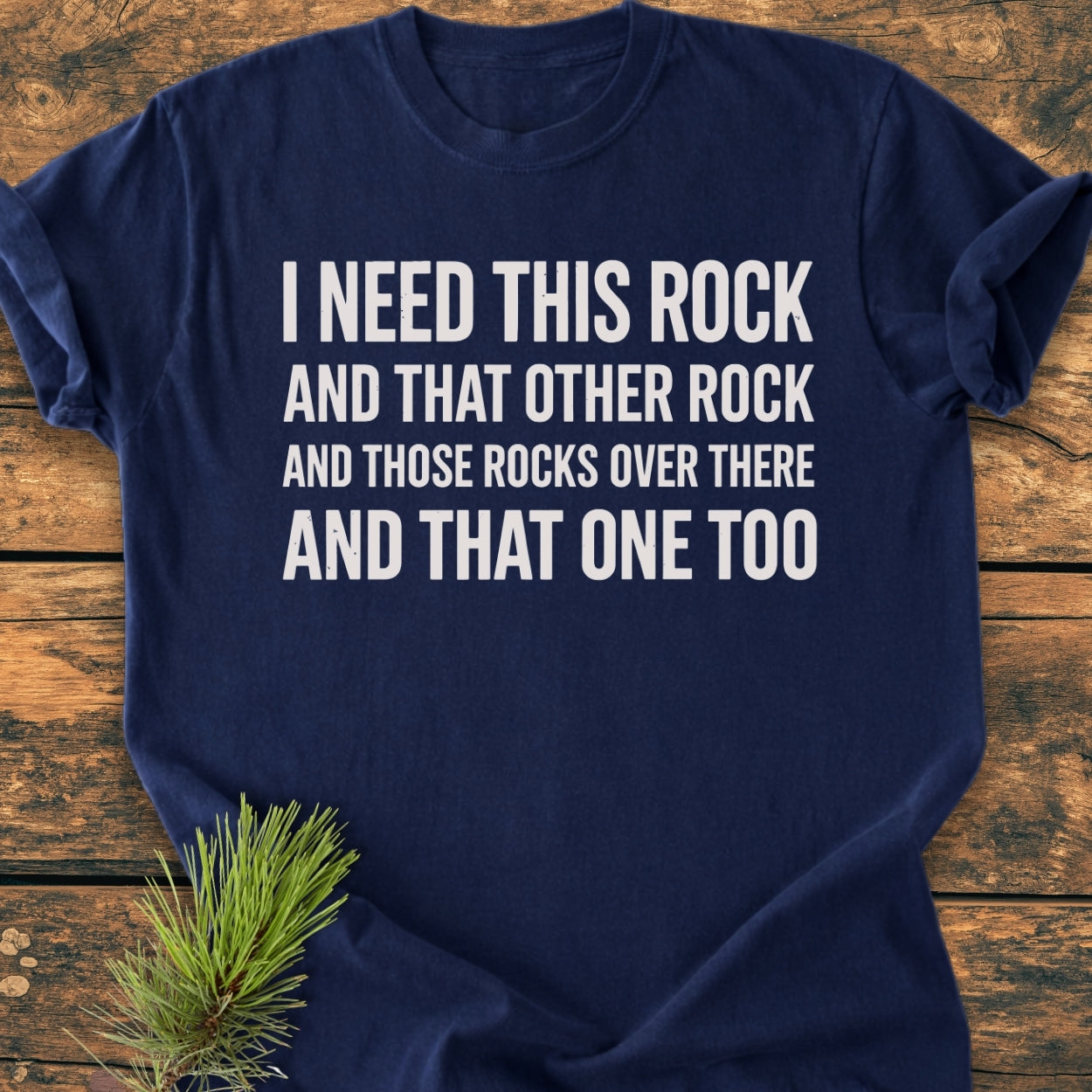 I Need This Rock - Vintage Wash Tee featuring white block text on a navy blue T-shirt, displayed on rustic wood with a pine sprig accent, emphasizing comfort and relaxed fit.