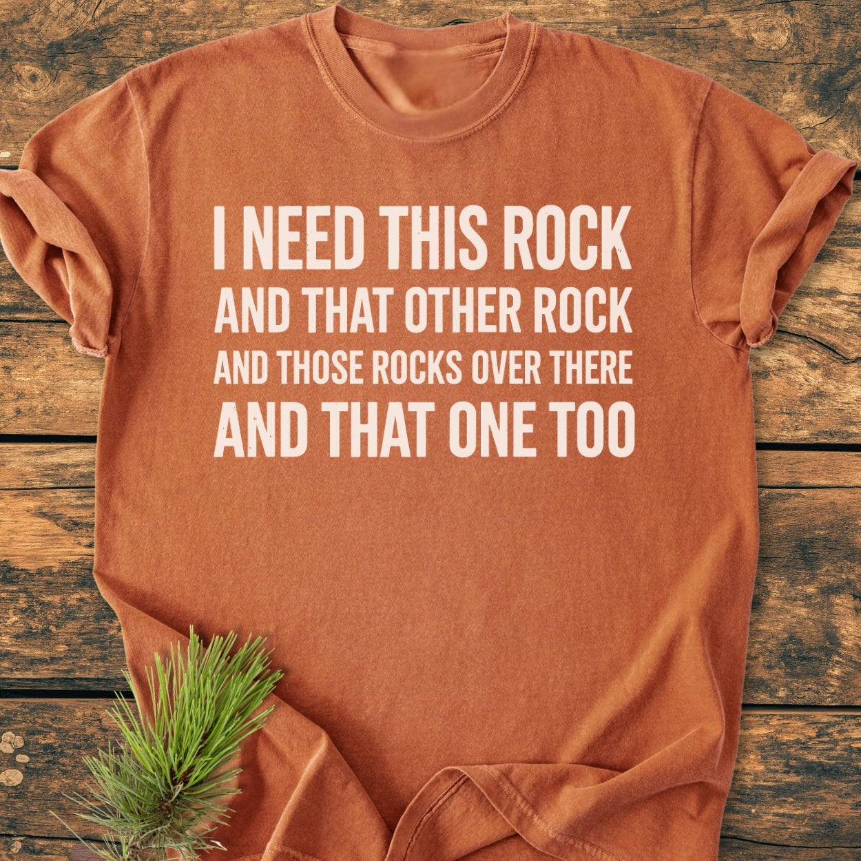 I Need This Rock - Vintage Wash Tee in rust-orange with a humorous rock-themed slogan, laid flat on wood, pine needles nearby, showcasing its relaxed fit and soft, durable fabric.