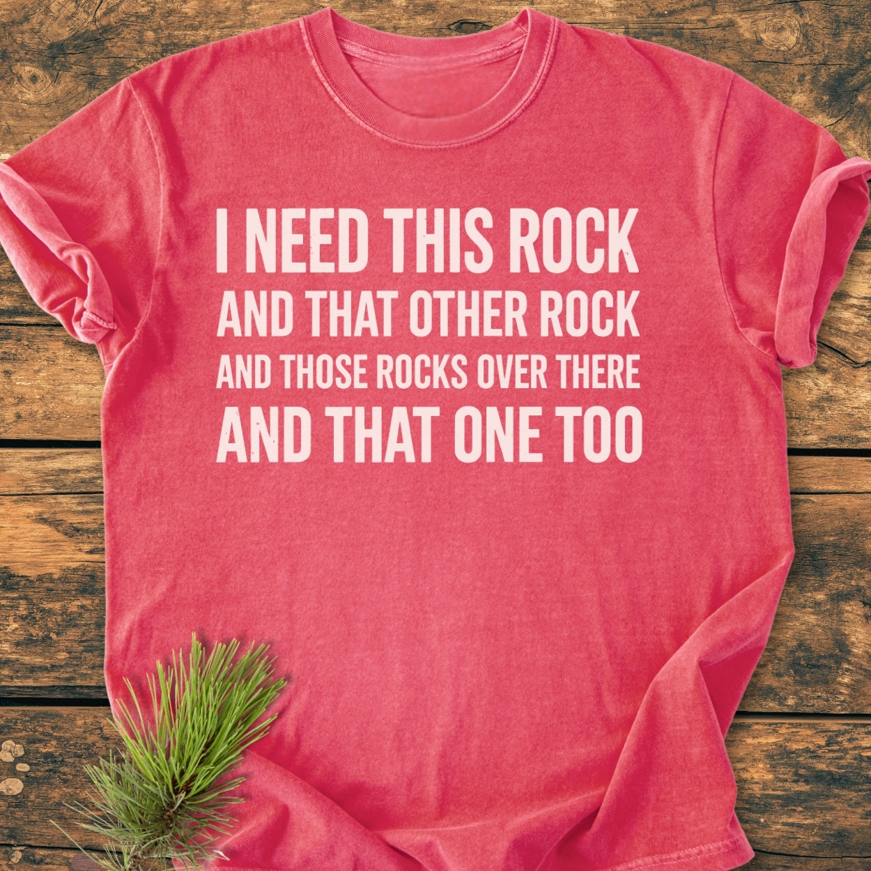 Heather-red I Need This Rock - Vintage Wash Tee with humorous rock-themed slogan in white, laid flat on rustic wood, featuring soft-washed, ring-spun cotton, with a relaxed fit, and durable double-needle stitching.