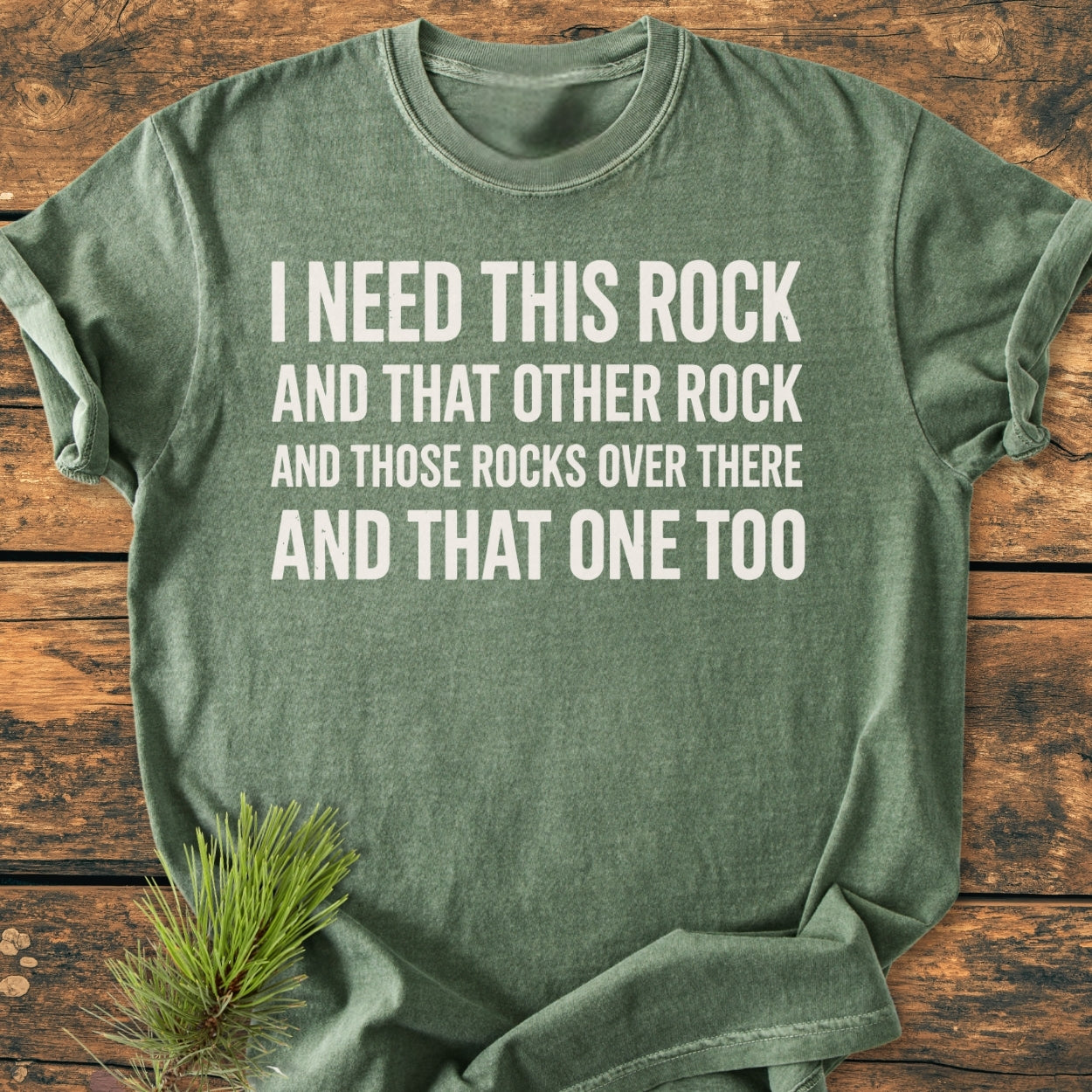 Heather-green I Need This Rock - Vintage Wash Tee with playful white text, laid on wood. Features include a relaxed fit and durable stitching. Accompanied by a pine sprig.