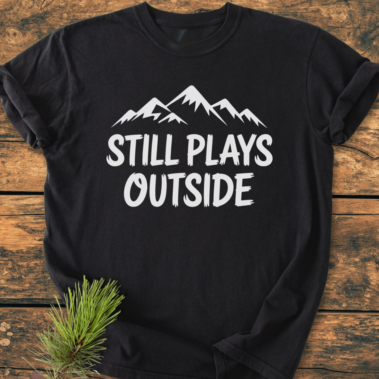 Still Plays Outside - Vintage Wash Tee features a white mountain graphic and slogan on a black T-shirt, laid flat with a pine sprig accent, showcasing its durable, relaxed fit.