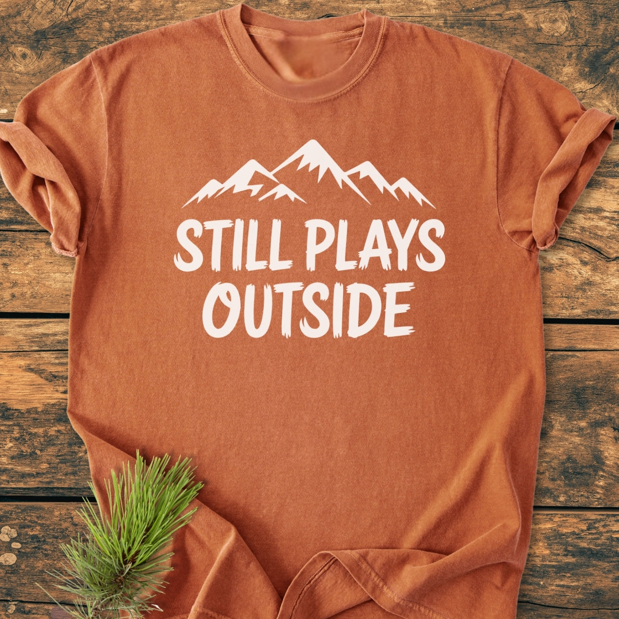 Still Plays Outside - Vintage Wash Tee in rust-orange features a white mountain silhouette and slogan, laid on wood with a pine sprig accent, showcasing its cozy, relaxed fit and durable construction.