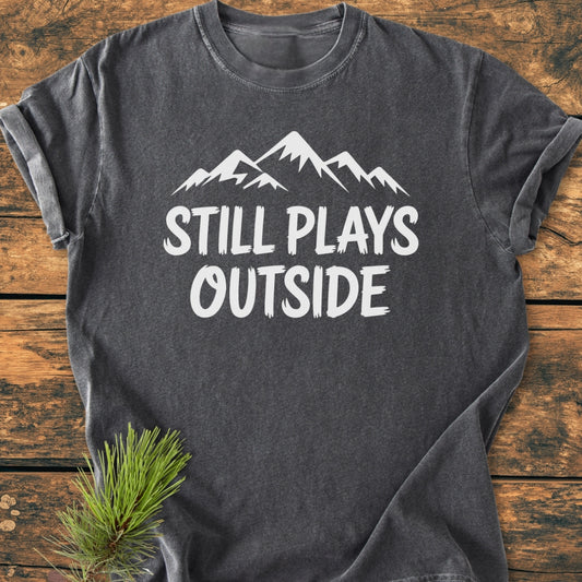 Still Plays Outside - Vintage Wash Tee features a white mountain graphic and phrase on a charcoal-gray shirt, styled with rolled sleeves and a pine branch. Relaxed fit and durable stitching.
