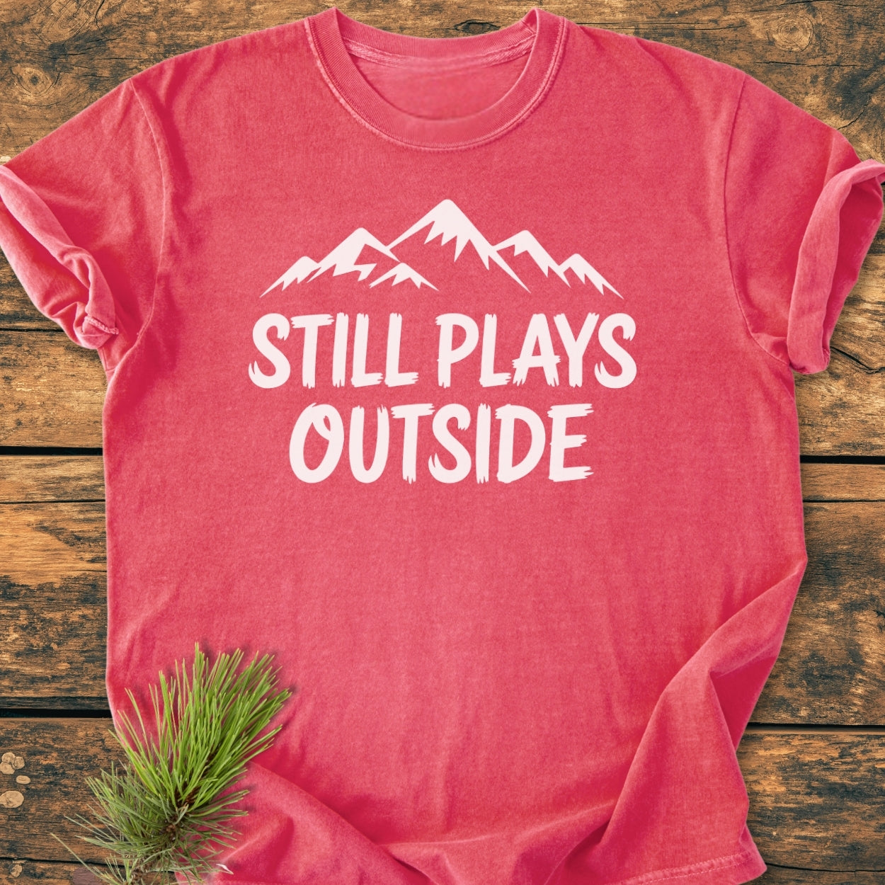 Still Plays Outside - Vintage Wash Tee in washed-red featuring a white mountain graphic and text, laid on rustic wood with a pine branch beside it, highlighting its outdoorsy appeal.