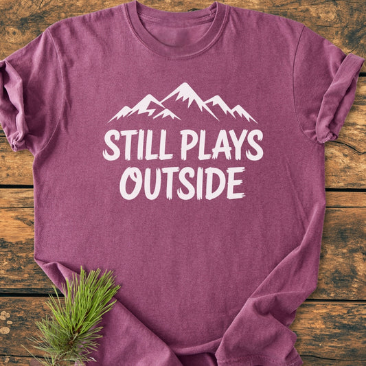 The Still Plays Outside - Vintage Wash Tee features a mountain silhouette and bold text on a heathered purple T-shirt, laid flat on rustic wood, with a pine sprig accentuating its outdoor theme.