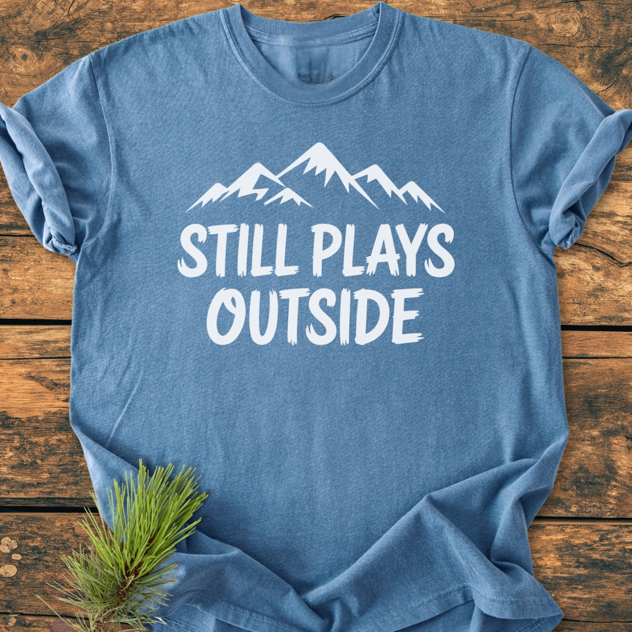 Still Plays Outside - Vintage Wash Tee features a heather-blue, rolled-sleeve design with a white mountain graphic and slogan, styled on a rustic surface, enhanced by a pine sprig accent.
