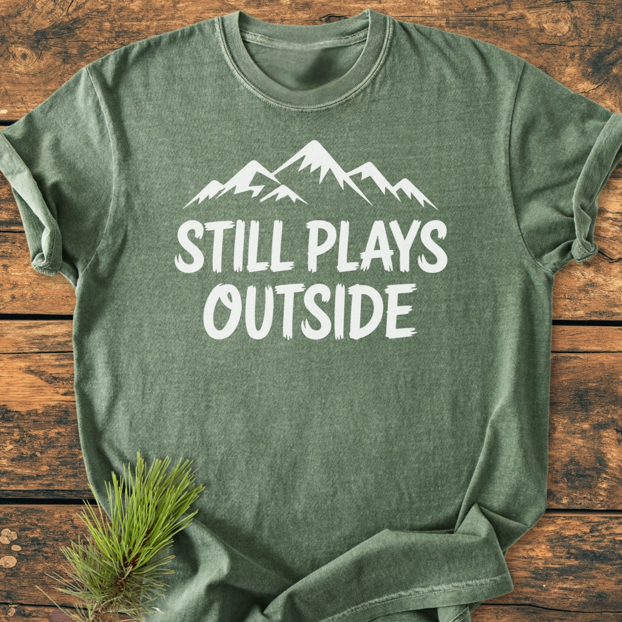 Green short-sleeved Still Plays Outside - Vintage Wash Tee with white mountain graphic and text on a wooden surface, featuring durable, soft-washed, garment-dyed fabric for a cozy, relaxed fit.