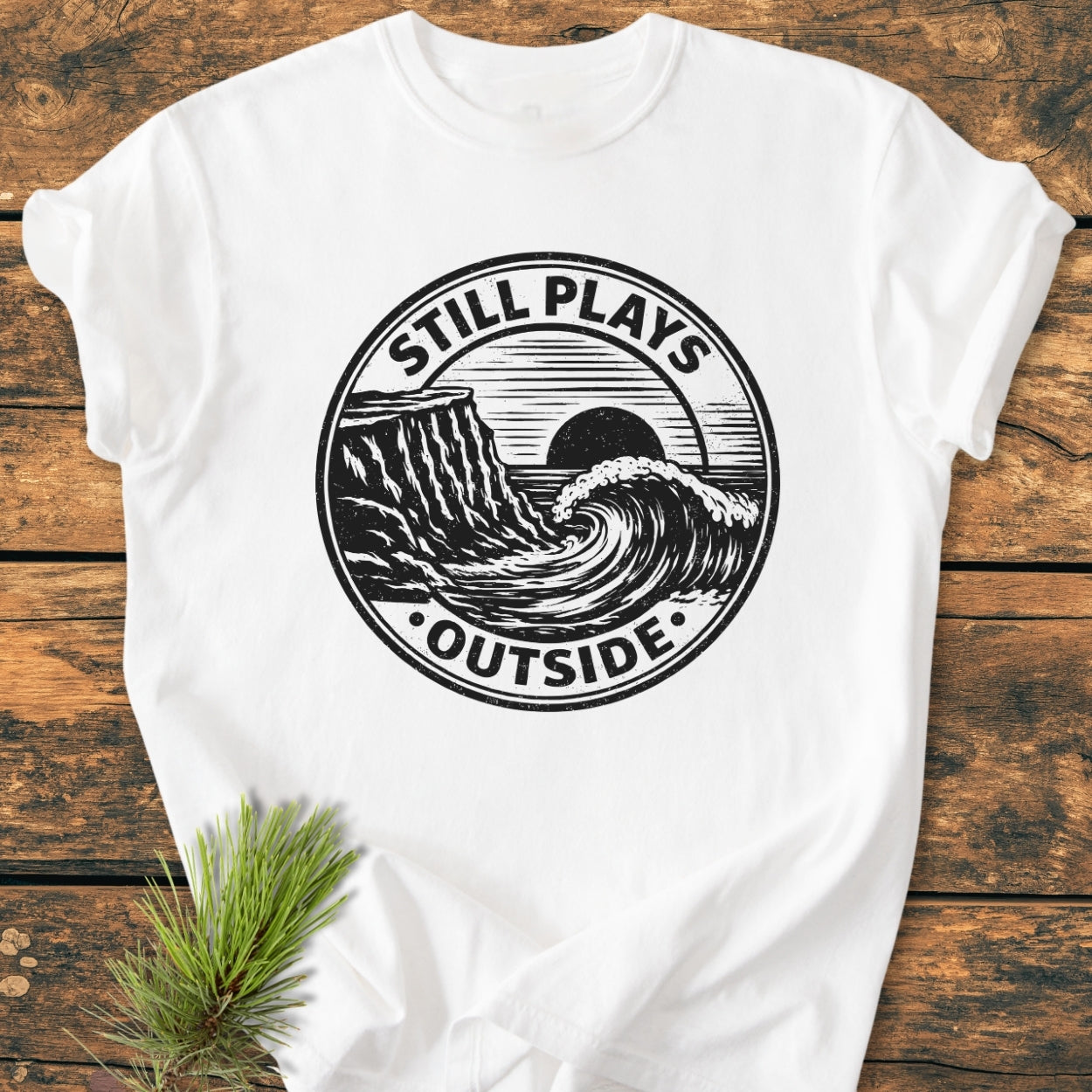 The Still Plays Outside - Vintage Wash Tee features a black graphic of a coastline and wave on a white t-shirt, laid flat with pine needles beside it, highlighting its relaxed, durable design.