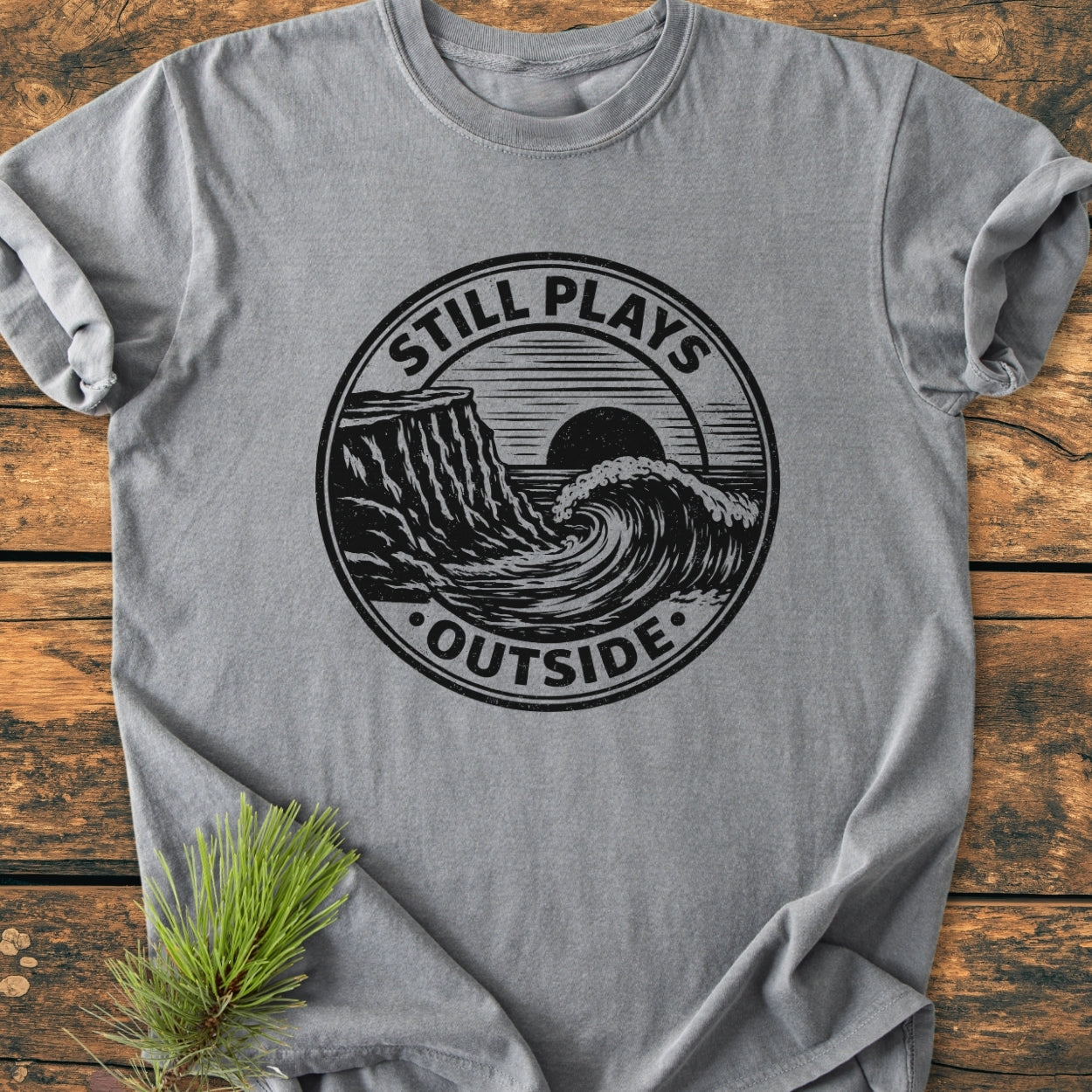 Still Plays Outside - Vintage Wash Tee features a black ocean wave graphic with sun and cliff on a heather-gray, soft-washed cotton shirt, laid on a wooden surface with a pine sprig.