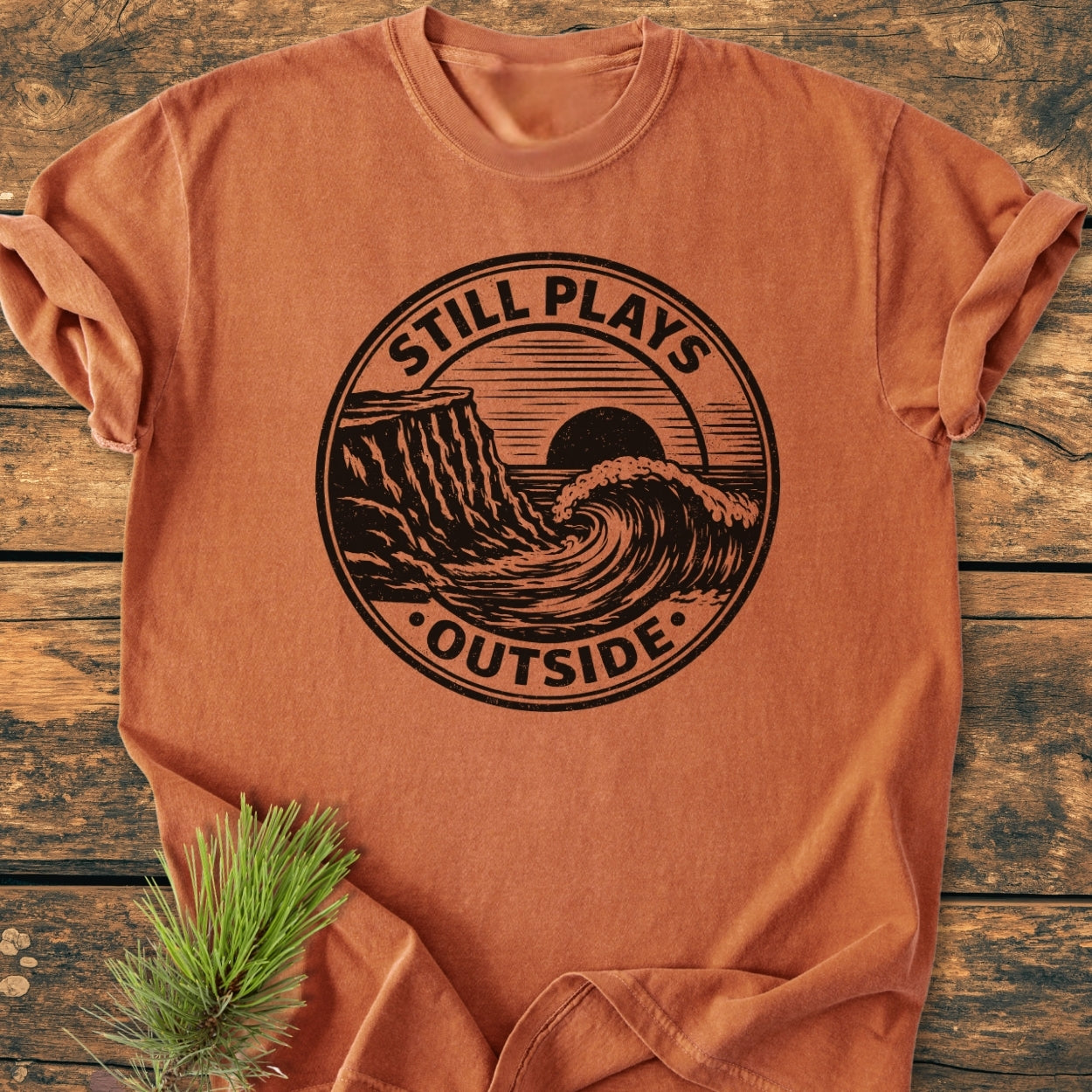 Still Plays Outside - Vintage Wash Tee featuring a black badge-style logo with a seaside cliff, wave, and sun, laid on wood with a pine sprig. Made from soft, durable, ring-spun cotton.