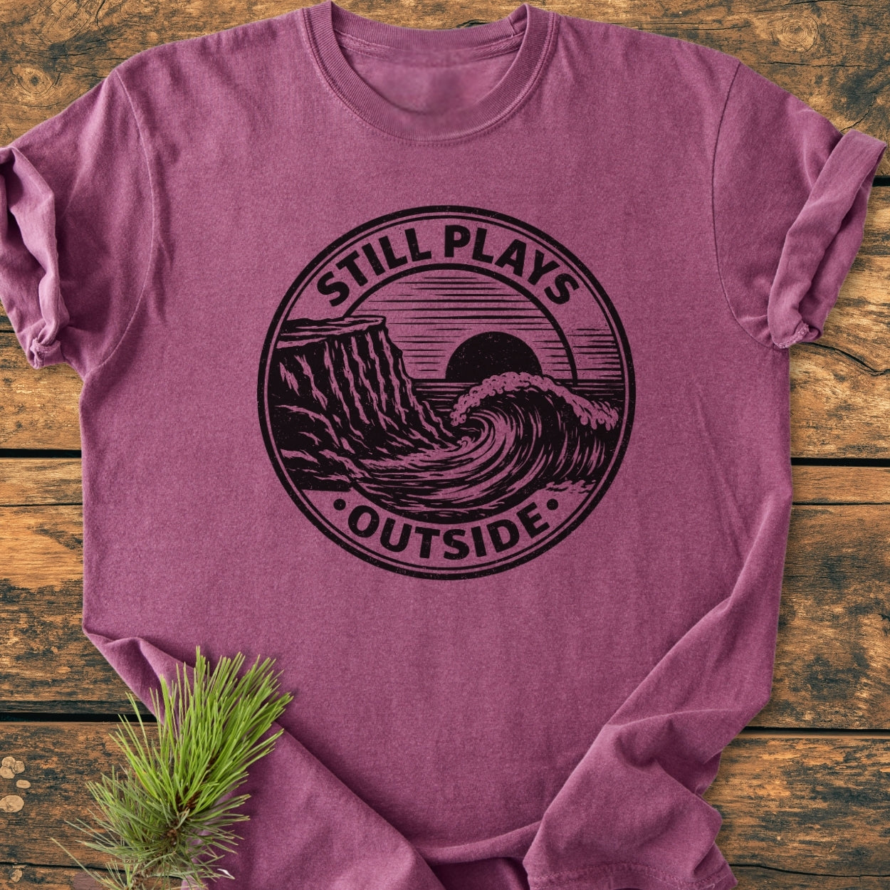 Still Plays Outside - Vintage Wash Tee features a circular coastline graphic on a heathered plum T-shirt, laid on wood, with a pine sprig beneath, highlighting its relaxed fit and durable stitching.