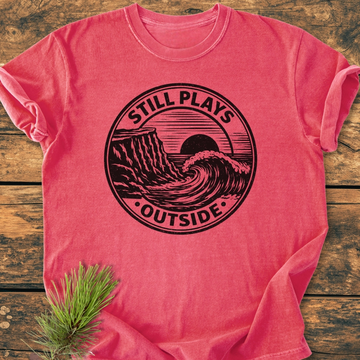 Still Plays Outside - Vintage Wash Tee on wooden surface, featuring a sun and wave design. Includes a pine sprig accent, highlighting the garment-dyed, relaxed fit style and durable stitching.