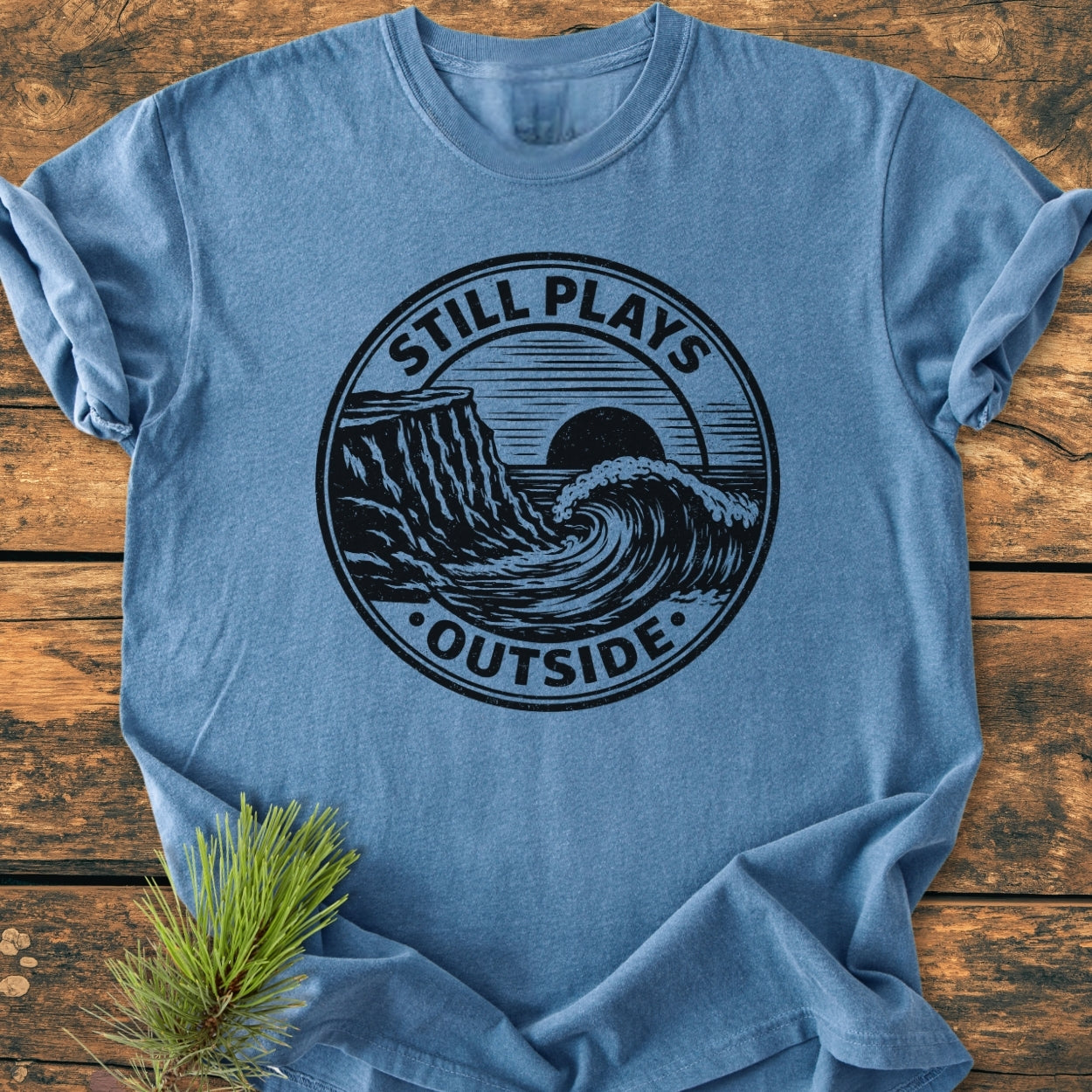 The Still Plays Outside - Vintage Wash Tee features a black graphic of a wave, cliff, and sun on the front, laid on wood with pine, showcasing its relaxed, durable design.