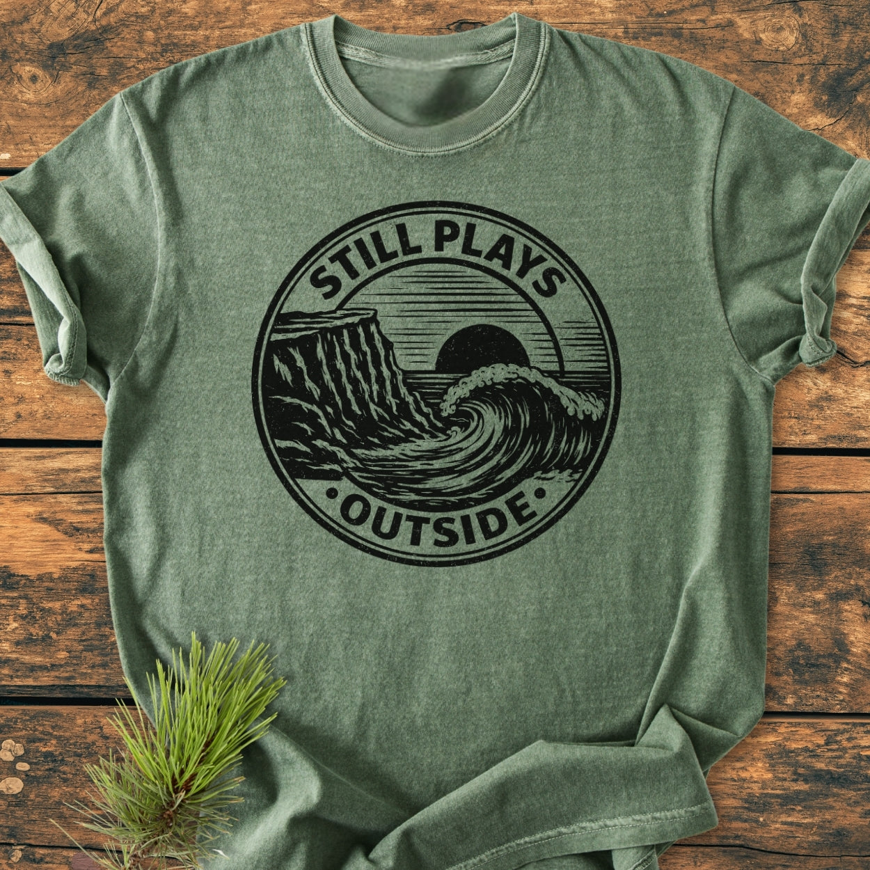 Still Plays Outside - Vintage Wash Tee in heather-green, featuring a black graphic with a cliff, wave, and sun. Displayed flat with a pine sprig accent. Made of soft, durable, garment-dyed cotton.