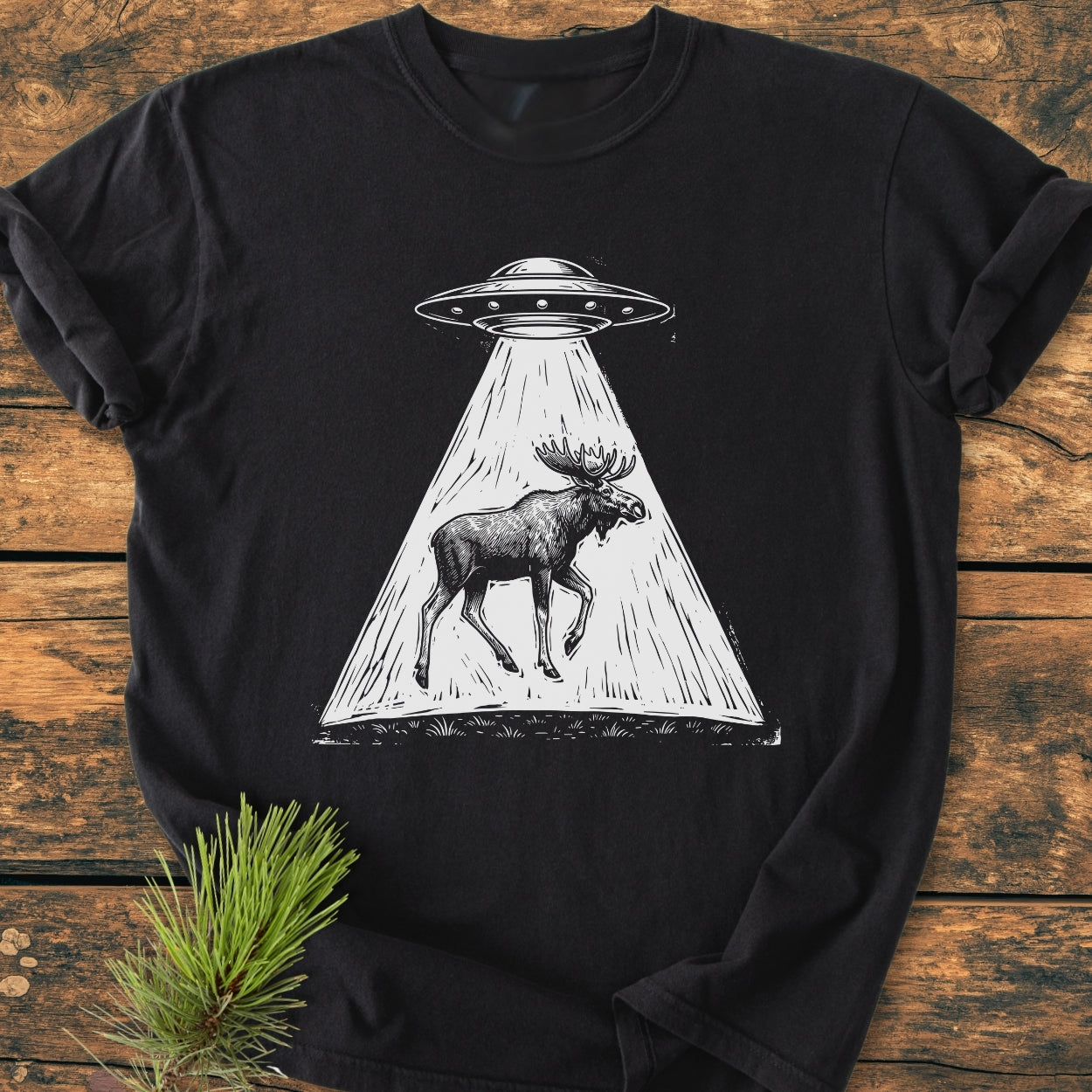 The Only Way Is Up - Vintage Wash Tee: Black T-shirt featuring a UFO beaming up a moose, laid on a wooden surface with a pine sprig beside it, emphasizing its cozy, relaxed fit and durable design.