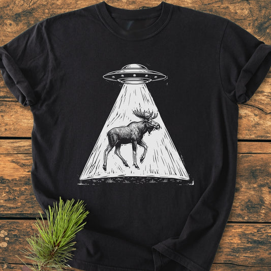 The Only Way Is Up - Vintage Wash Tee: Black T-shirt featuring a UFO beaming up a moose, laid on a wooden surface with a pine sprig beside it, emphasizing its cozy, relaxed fit and durable design.