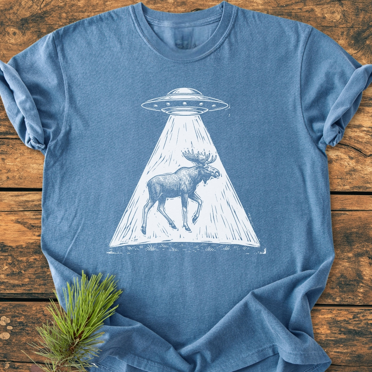 The Only Way Is Up - Vintage Wash Tee features a white line-drawing of a UFO beaming up a moose, laid out on a wooden surface with a pine bough beside it.