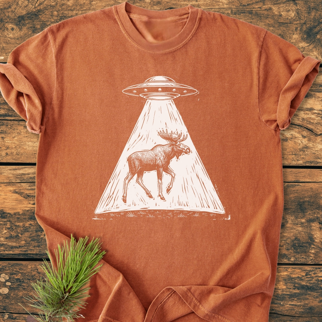 The Only Way Is Up - Vintage Wash Tee: A burnt-orange T-shirt featuring a white line-drawing of a flying-saucer UFO beaming up a moose, displayed on weathered wooden boards with a pine sprig accent.