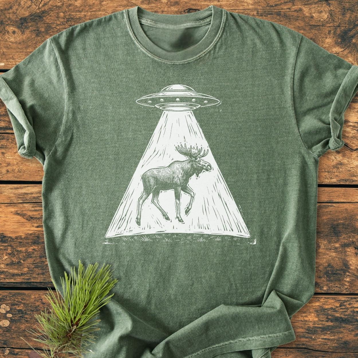 The Only Way Is Up - Vintage Wash Tee features an olive-green T-shirt with a white flying saucer graphic beaming up a moose, laid on wood with pine sprig.