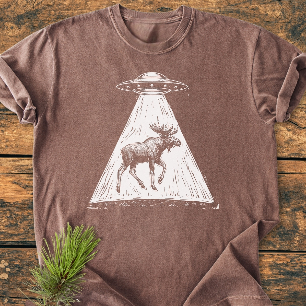 The Only Way Is Up - Vintage Wash Tee features a white UFO and moose graphic on a brown, relaxed-fit Comfort Colors t-shirt, styled with a pine sprig on a wooden surface.