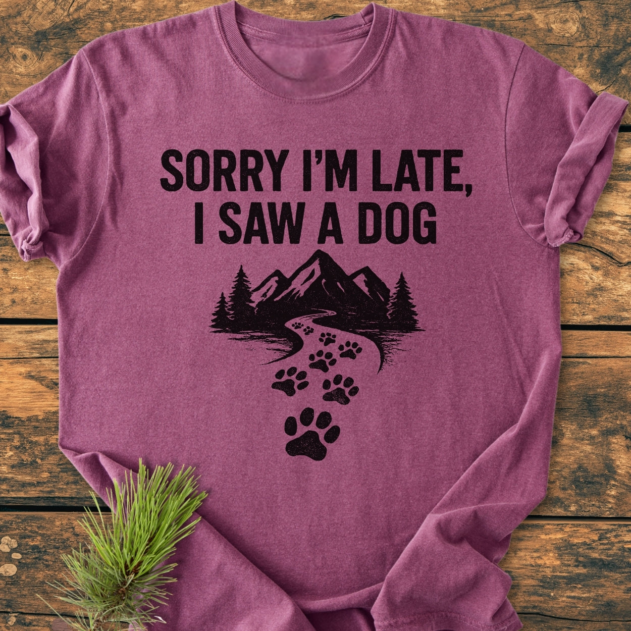Sorry I'm Late - Vintage Wash Tee in heather-purple, featuring bold black text and a graphic of paw prints forming a trail through pine trees and mountains, laid on a wooden surface.
