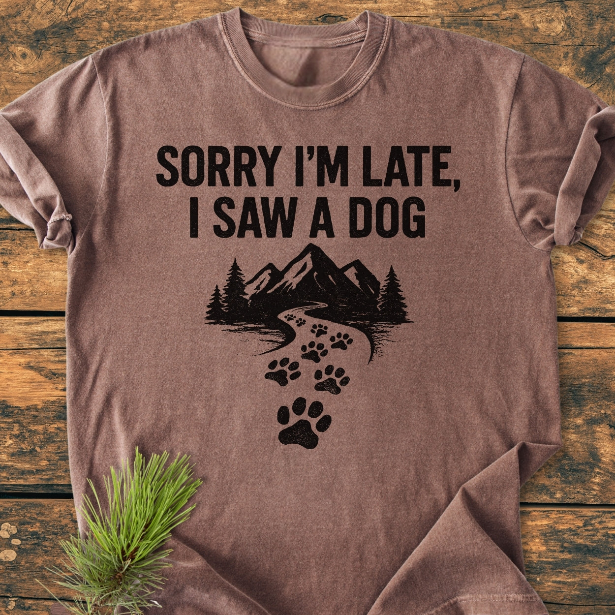 The Sorry I'm Late - Vintage Wash Tee features a playful dog-themed print and nature elements on a laid-flat brown T-shirt, highlighting its cozy, relaxed fit on a rustic wood surface.