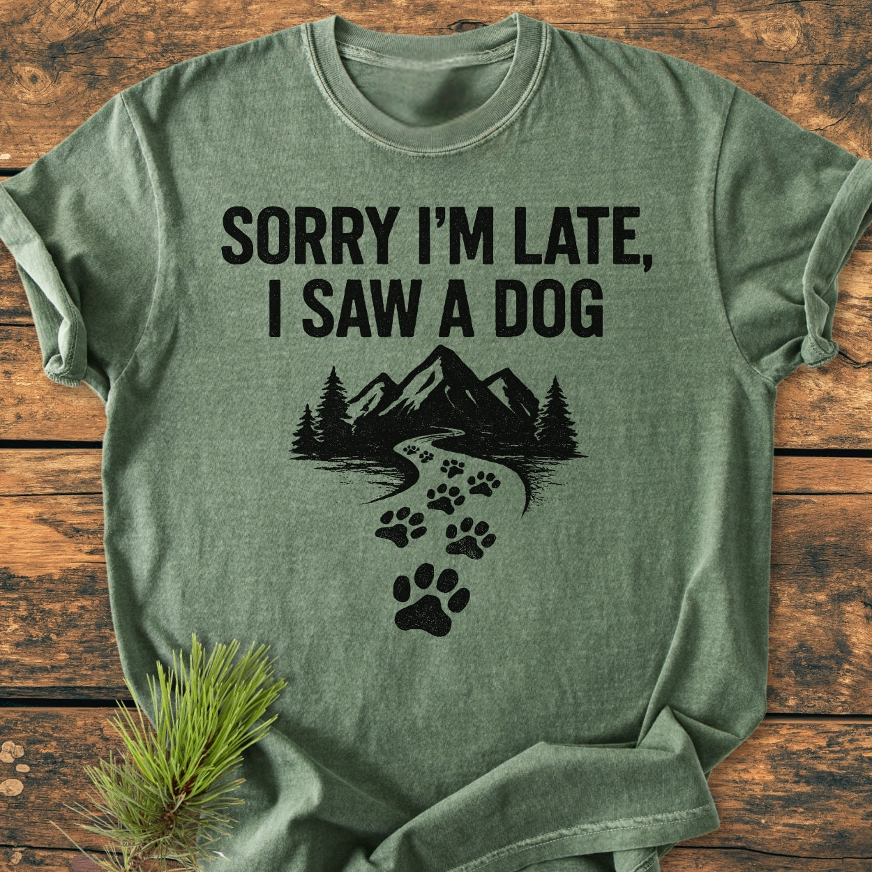 Sorry I'm Late - Vintage Wash Tee features a bold dog-themed graphic with mountains and paw prints, laid on wood with pine sprig, highlighting its relaxed fit and durable, soft-washed cotton fabric.