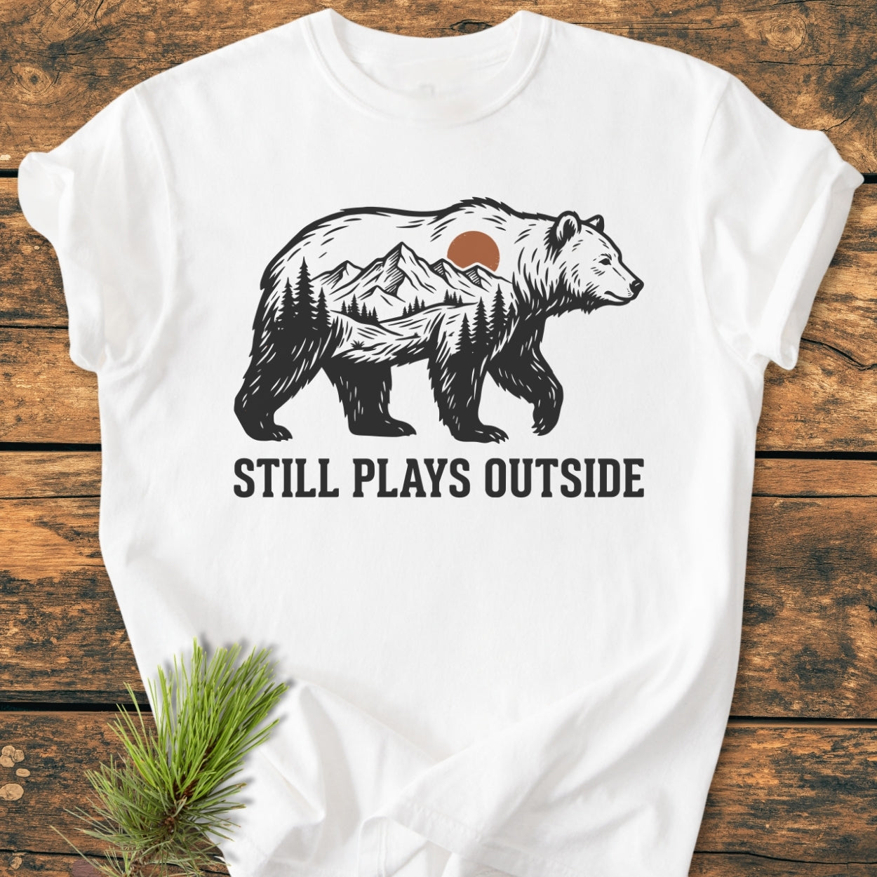Still Plays Outside - Vintage Wash Tee featuring a bear silhouette with a mountain scene, laid on wood with a pine twig beside. Made from soft, durable 100% ring-spun cotton for everyday comfort.