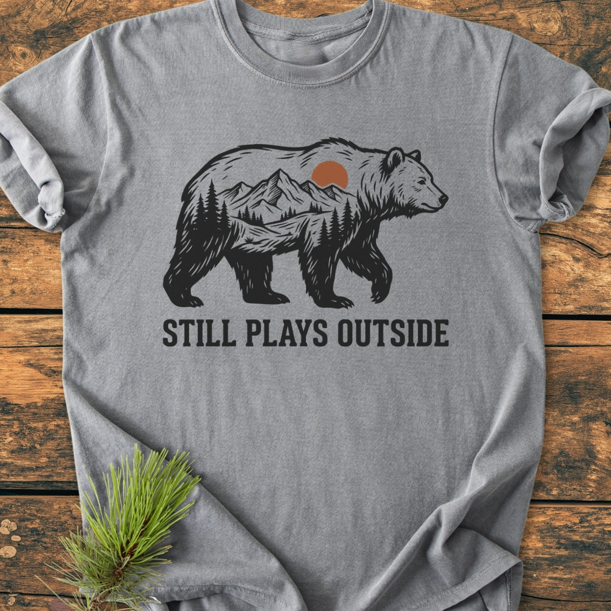 Still Plays Outside - Vintage Wash Tee: Gray t-shirt with a black bear silhouette featuring a mountain scene, laid flat on wood. Comfortable, relaxed fit, durable ring-spun cotton, perfect for outdoor enthusiasts.