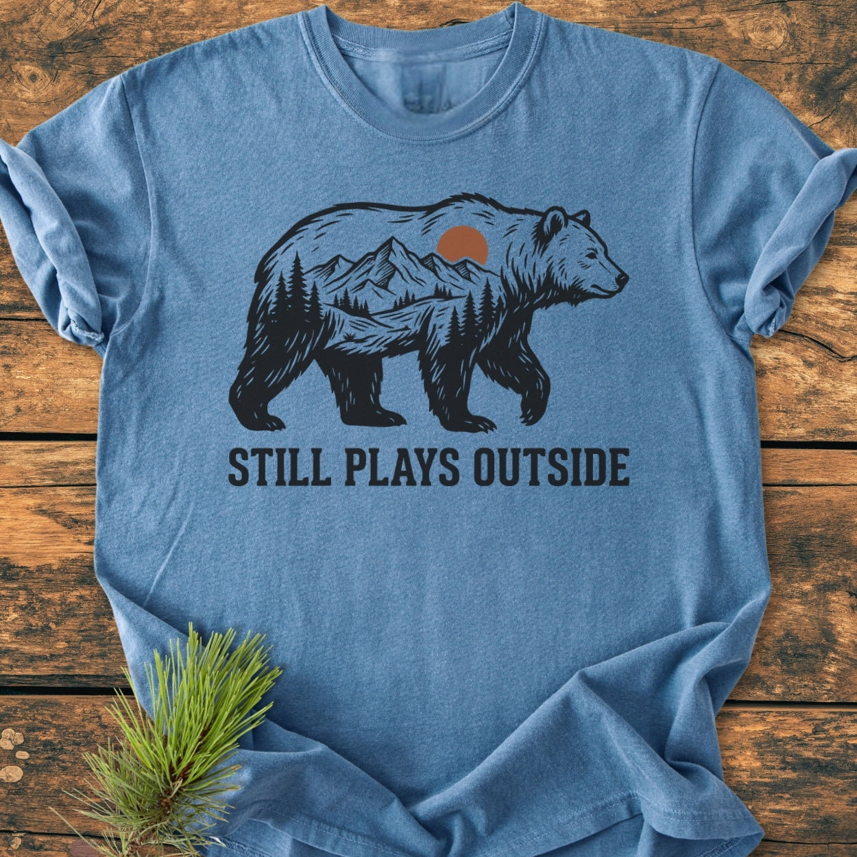 Heather-blue Still Plays Outside - Vintage Wash Tee featuring a bear silhouette with a mountain scene, laid on wood. Pine sprig beside it, showcasing the tee's relaxed fit and garment-dyed softness.