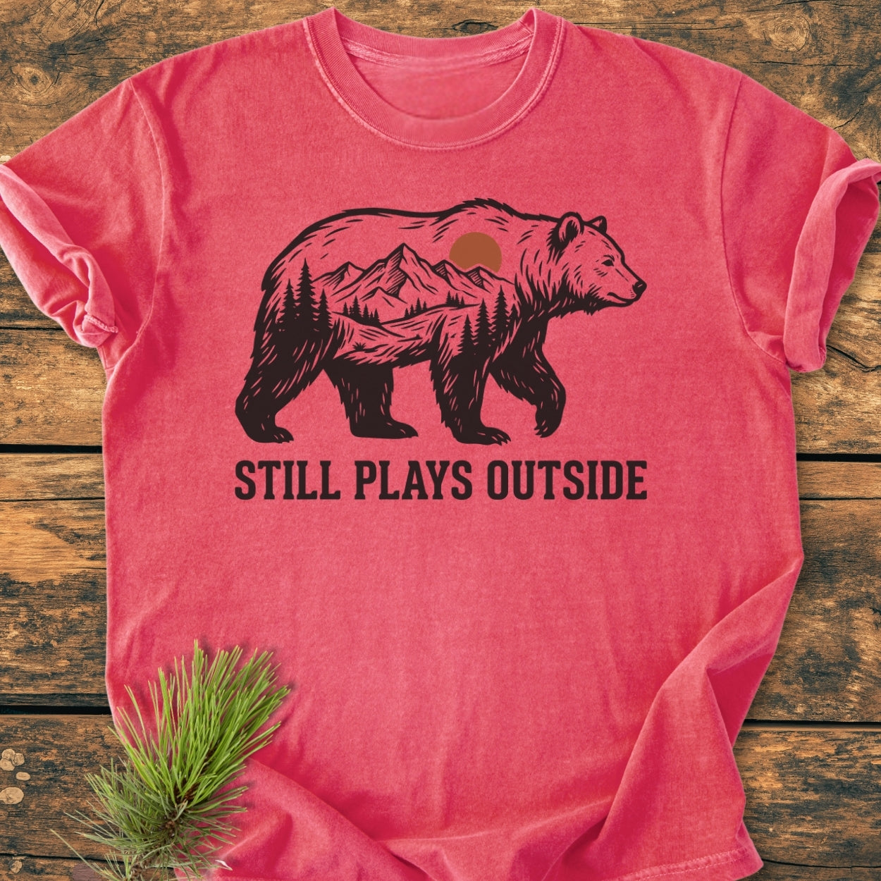 Still Plays Outside - Vintage Wash Tee featuring a stylized bear with mountain scene, on a heathered coral-red background, laid on wood with a pine sprig. Comfortable, durable, and relaxed fit.