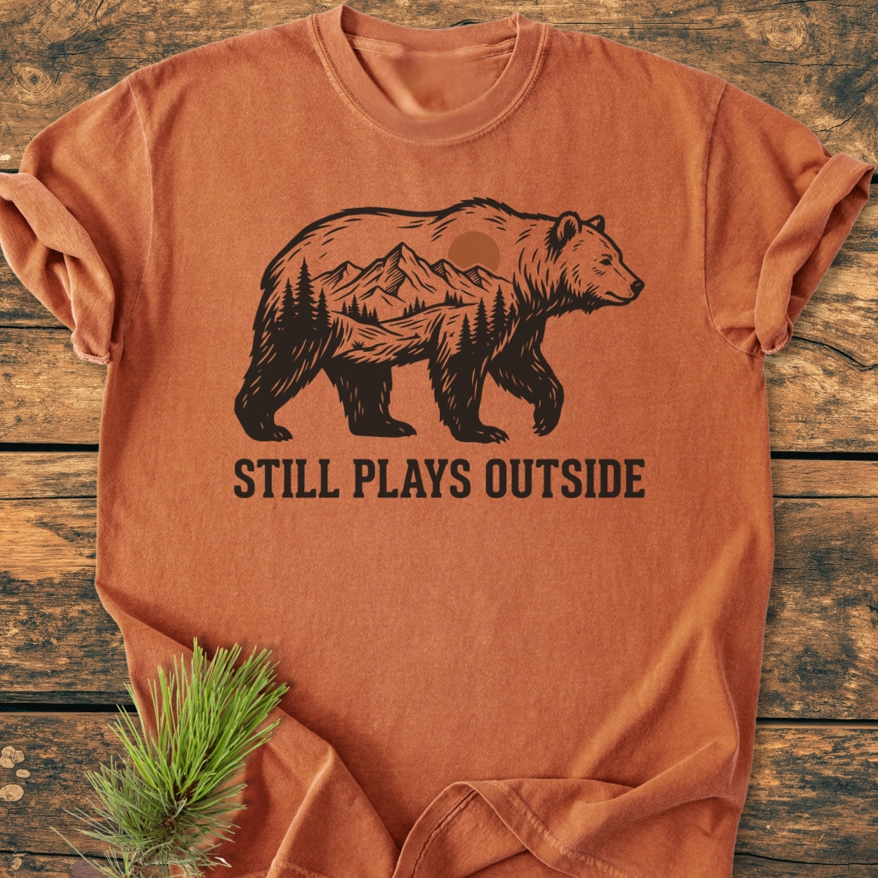 Still Plays Outside - Vintage Wash Tee featuring a black bear silhouette with a forest scene on rust-orange fabric, laid on wood. Includes text STILL PLAYS OUTSIDE beneath graphic. Pine sprig beside.