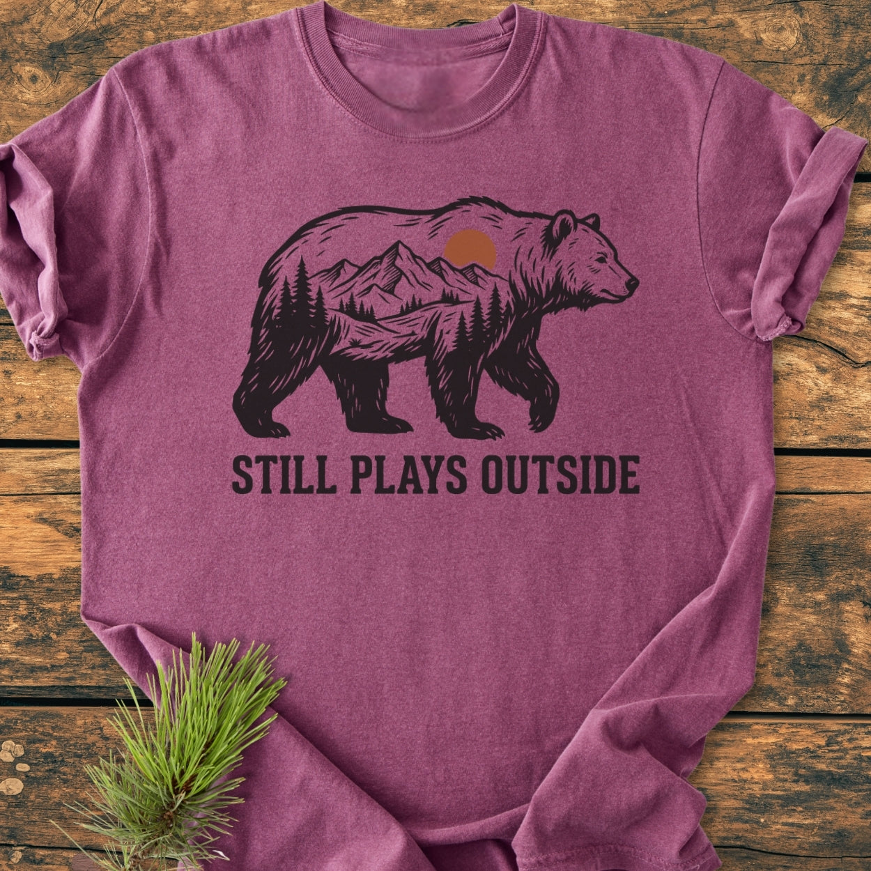 Still Plays Outside - Vintage Wash Tee with a bear silhouette featuring mountains and trees, laid on wood. Includes a pine bough as a prop, emphasizing its outdoor theme and relaxed fit.
