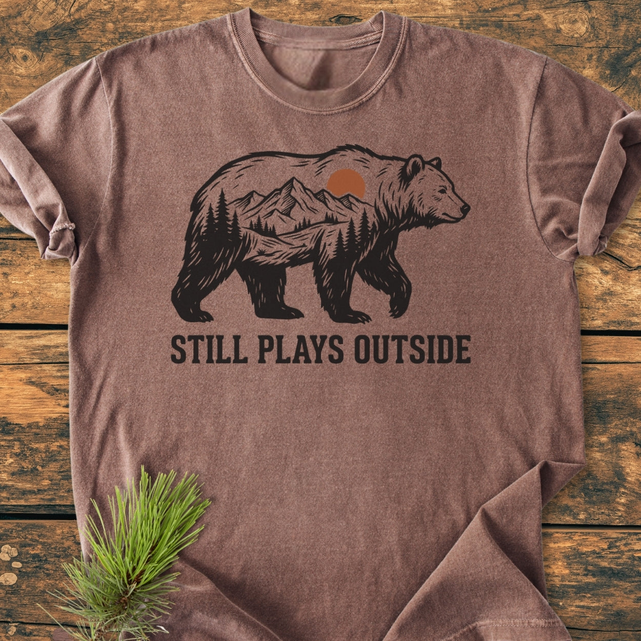 Still Plays Outside - Vintage Wash Tee featuring a bear silhouette with a mountain scene, laid on wood. Pine sprig accentuates its outdoorsy theme, highlighting the tee's relaxed fit and soft-washed fabric.