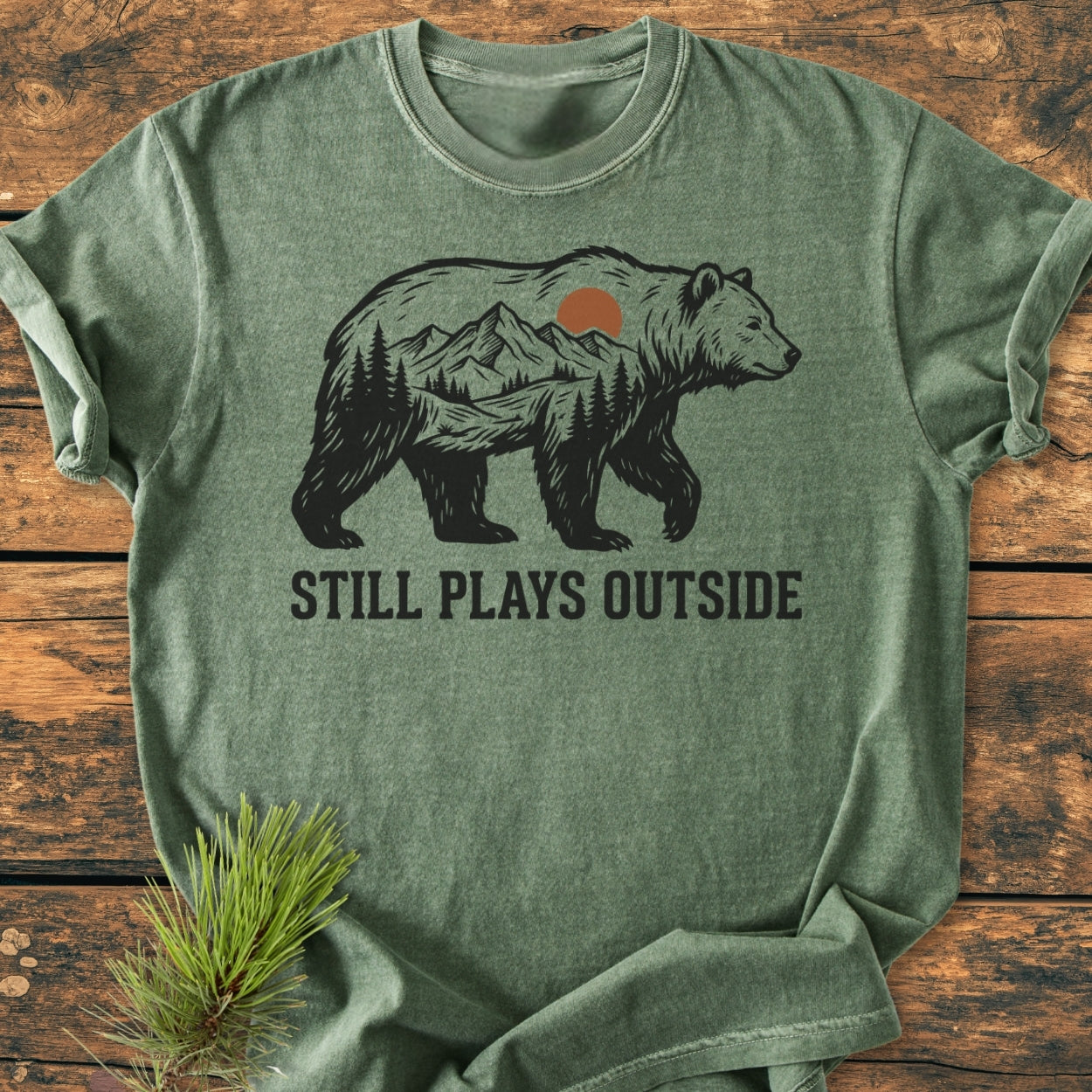 Heather-green Still Plays Outside - Vintage Wash Tee featuring a bear silhouette with a mountain scene, laid flat on wood. Includes bold text and pine branch detail, highlighting its relaxed fit and soft texture.