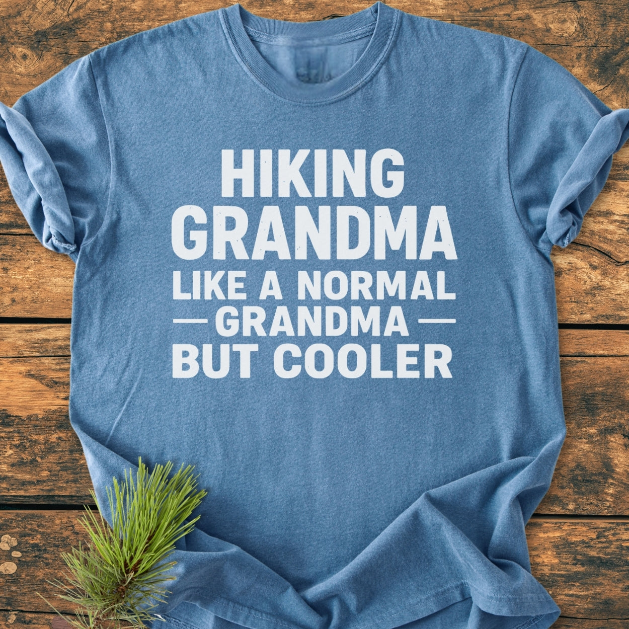 Heather-blue Hiking Grandma - Vintage Wash Tee with bold white slogan, laid on wood surface; pine sprig accent. Features relaxed fit, durable double-needle stitching, soft-washed 100% ring-spun cotton fabric.