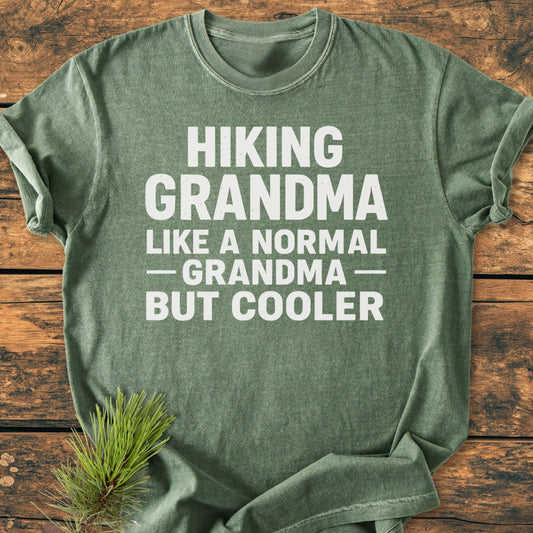 Heather-green Hiking Grandma - Vintage Wash Tee with playful slogan in white, laid flat on wood. Features soft, garment-dyed cotton, relaxed fit, and durable double-needle stitching. Pine sprig accentuates design.