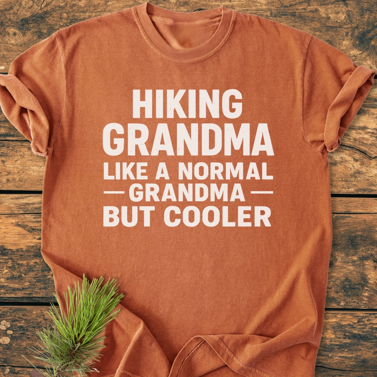 Burnt-orange Hiking Grandma - Vintage Wash Tee folded on wood, featuring a white slogan. Made from soft, garment-dyed, 100% ring-spun cotton, it offers a relaxed fit and durable stitching.