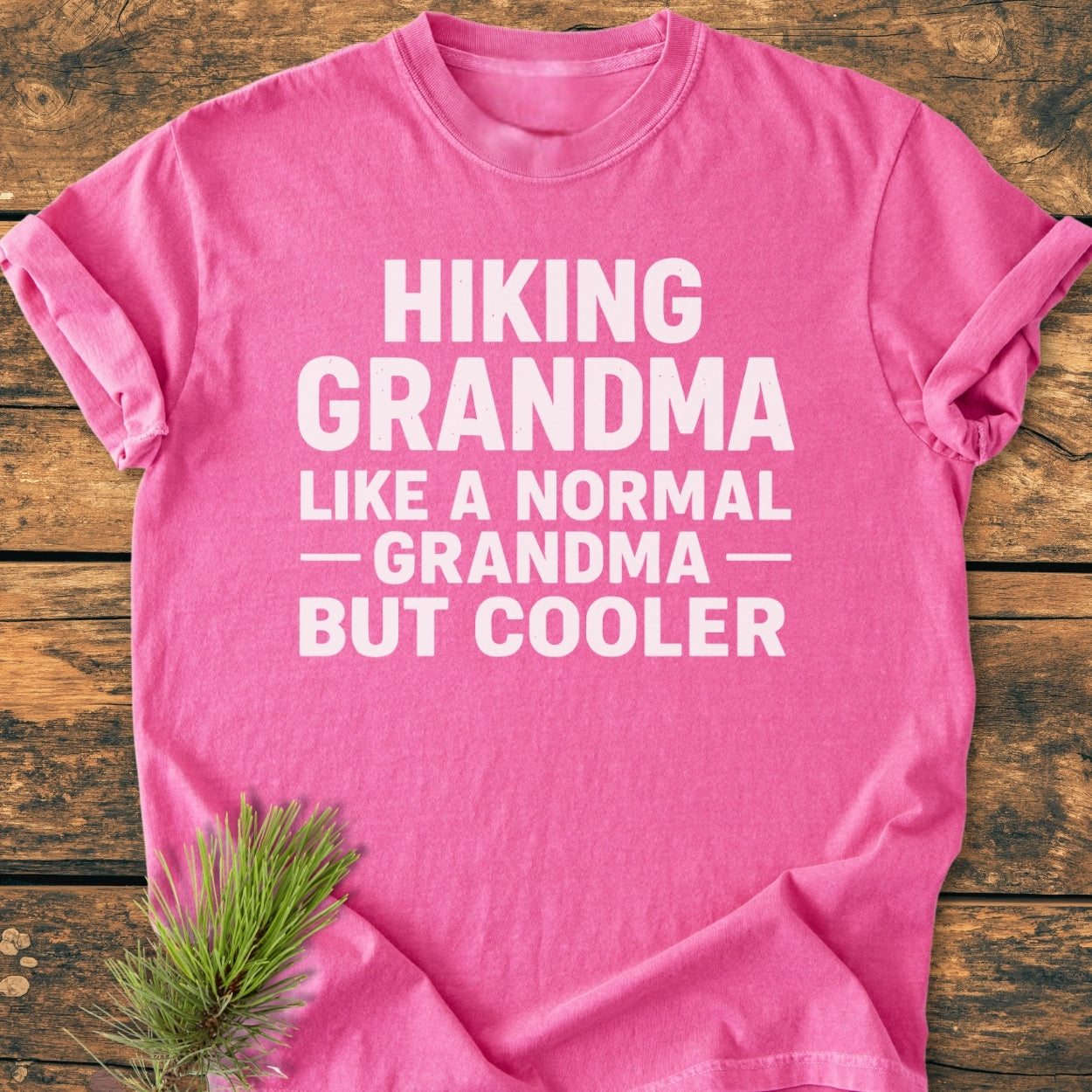 Pink Hiking Grandma - Vintage Wash Tee with bold white slogan text, laid on wood. Features pine sprig, highlighting the soft-washed, relaxed fit, 100% ring-spun cotton design.