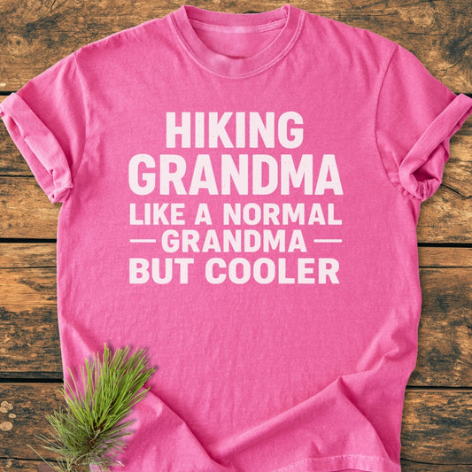 Pink Hiking Grandma - Vintage Wash Tee with bold white slogan text, laid on wood. Features pine sprig, highlighting the soft-washed, relaxed fit, 100% ring-spun cotton design.