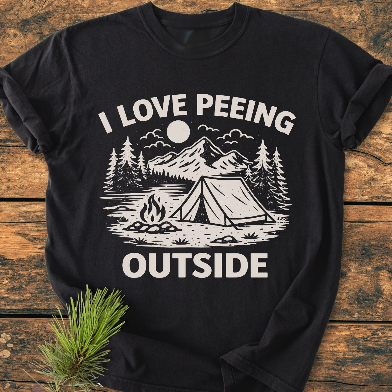 I Love Peeing Outside - Vintage Wash Tee features a black T-shirt with a white camping scene and humorous slogan, laid flat on wood, highlighting its relaxed fit and durable design.