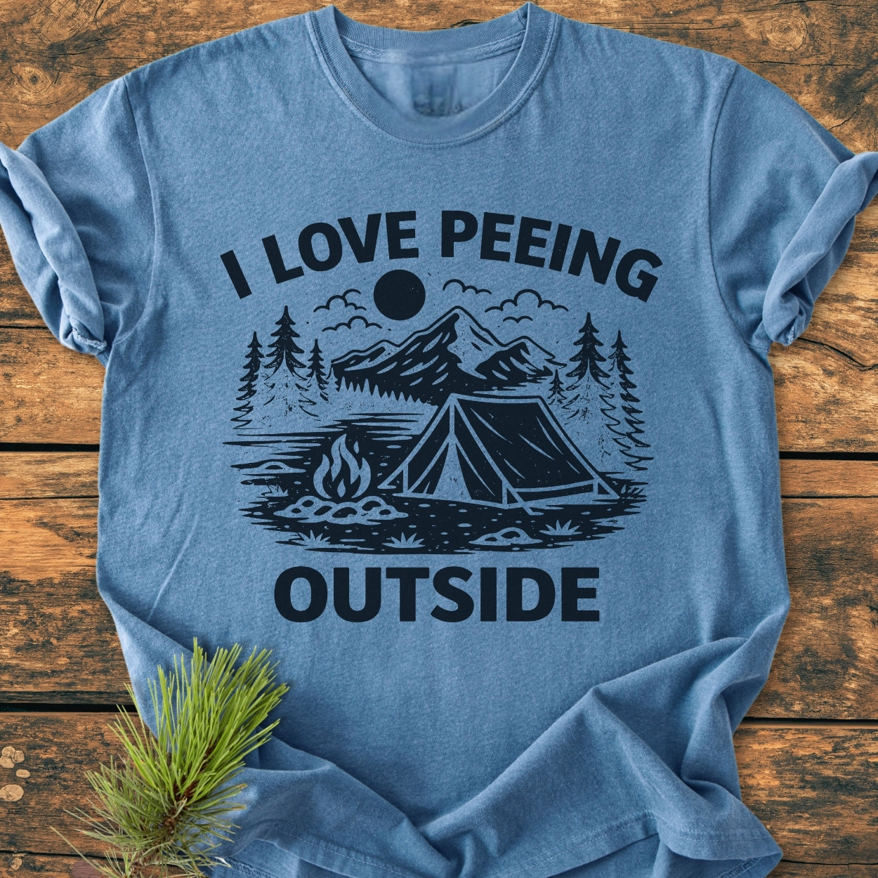 I Love Peeing Outside - Vintage Wash Tee features a light-blue design with a stylized camping scene and cheeky slogan, laid on wood with a pine branch beside it, showcasing its relaxed fit and durability.