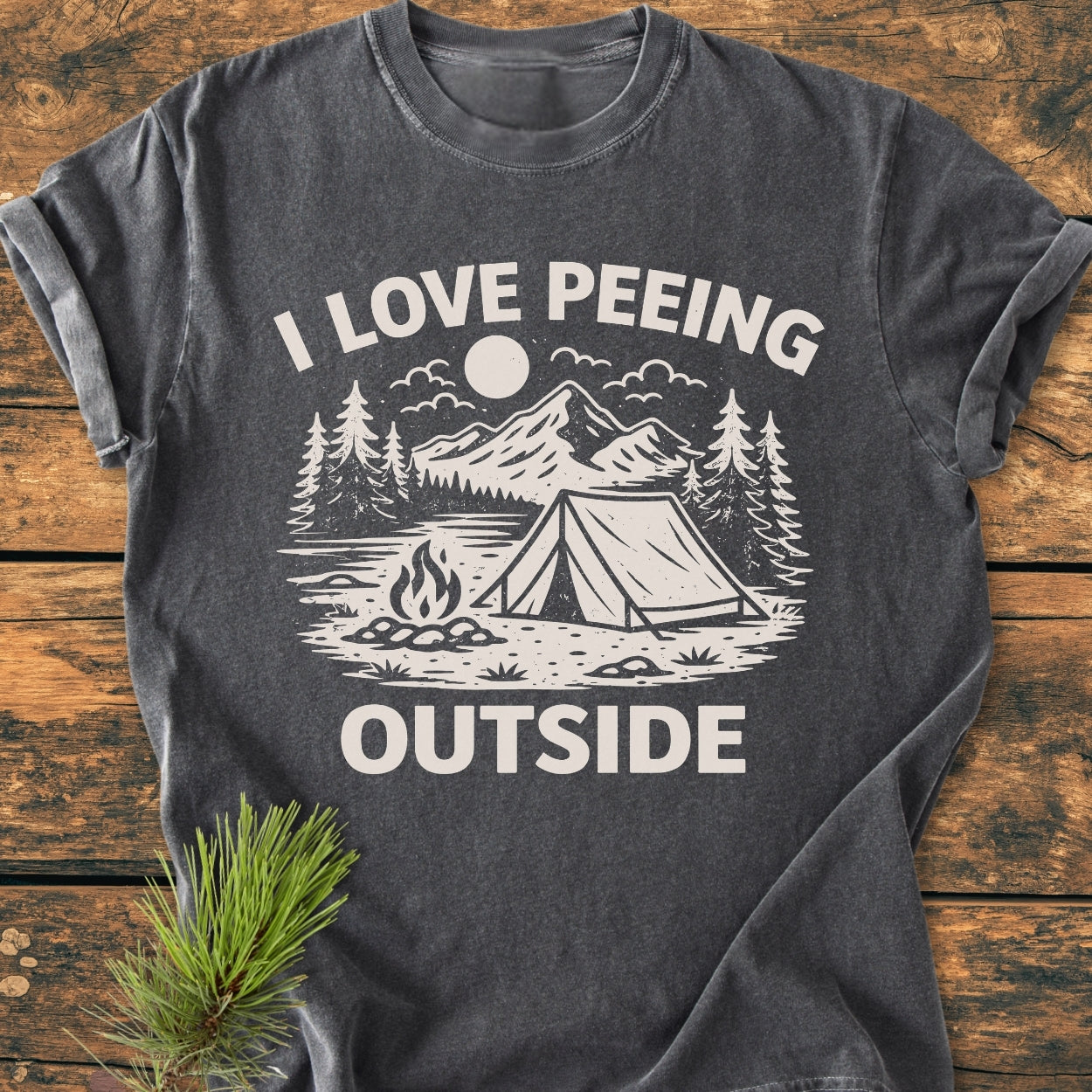 The I Love Peeing Outside - Vintage Wash Tee showcases a camping scene with mountains, trees, and a tent, printed on a dark gray T-shirt, emphasizing comfort and durability, accented with a pine sprig.