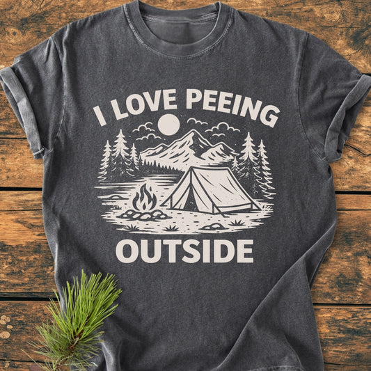 The I Love Peeing Outside - Vintage Wash Tee showcases a camping scene with mountains, trees, and a tent, printed on a dark gray T-shirt, emphasizing comfort and durability, accented with a pine sprig.