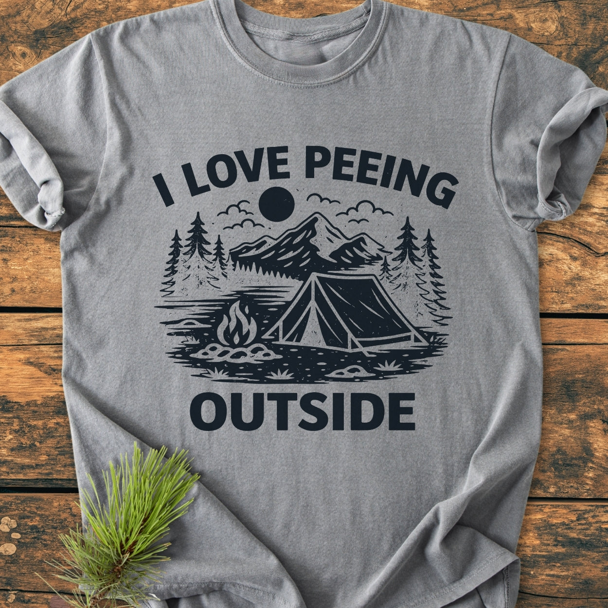 I Love Peeing Outside - Vintage Wash Tee featuring a stylized camping scene with pine trees and mountains, laid on wood with a pine sprig, highlighting the tee's relaxed fit and durable design.
